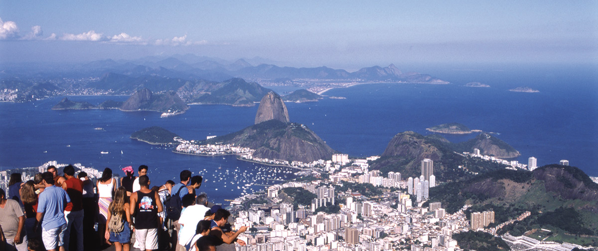 Aerial view of Rio de Janeiro's cityscape from a scenic viewpoint, showing Sugarloaf Mountain, the harbor, and city buildings spread along the coastline, with tourists observing from a viewing platform.