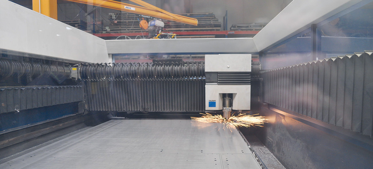 Industrial laser cutting machine in operation, showing sparks flying inside a safety enclosure with black protective curtains and overhead crane system.