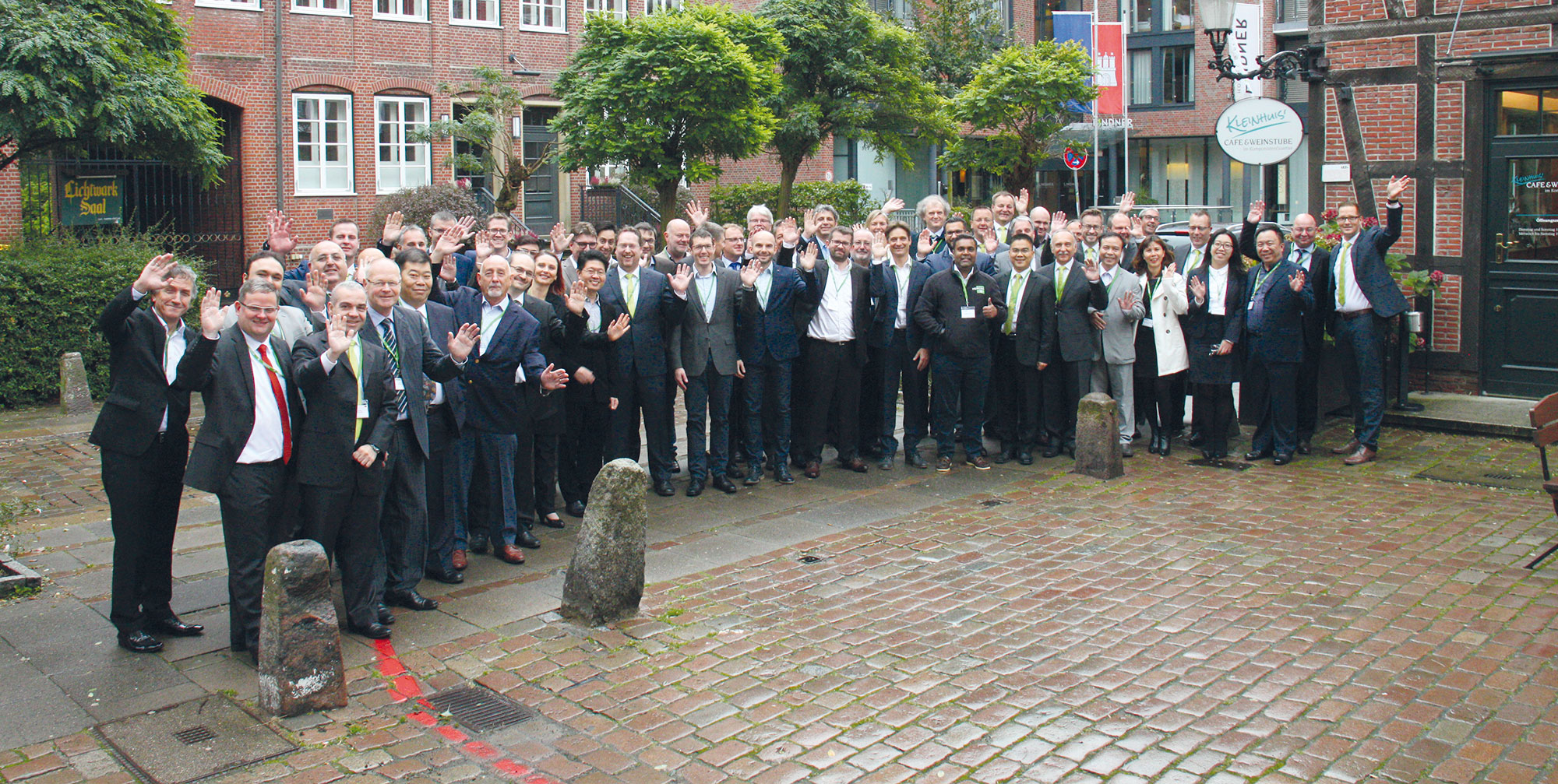 Large group of business professionals in suits waving at camera while standing in front of brick building with traditional architecture