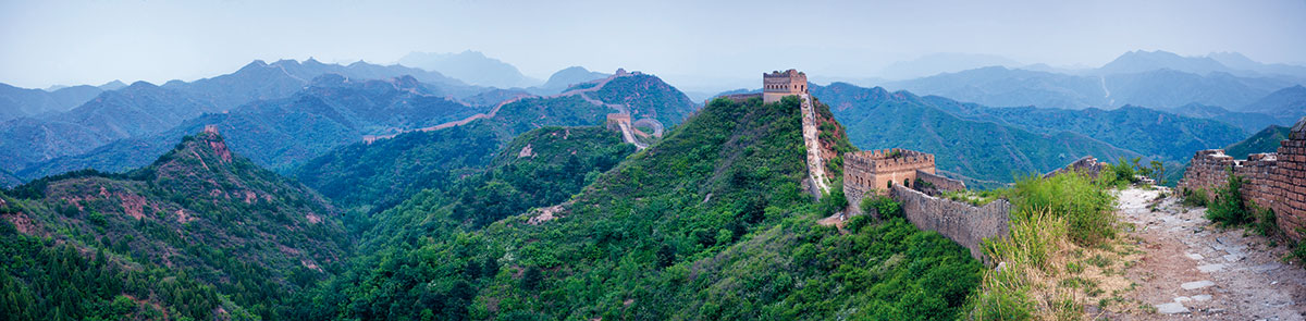 Chinesische Mauer schlängelt sich durch grüne Berglandschaft, Panoramablick auf historisches Bauwerk im Morgendunst mit Wachtürmen und bergiger Horizontlinie