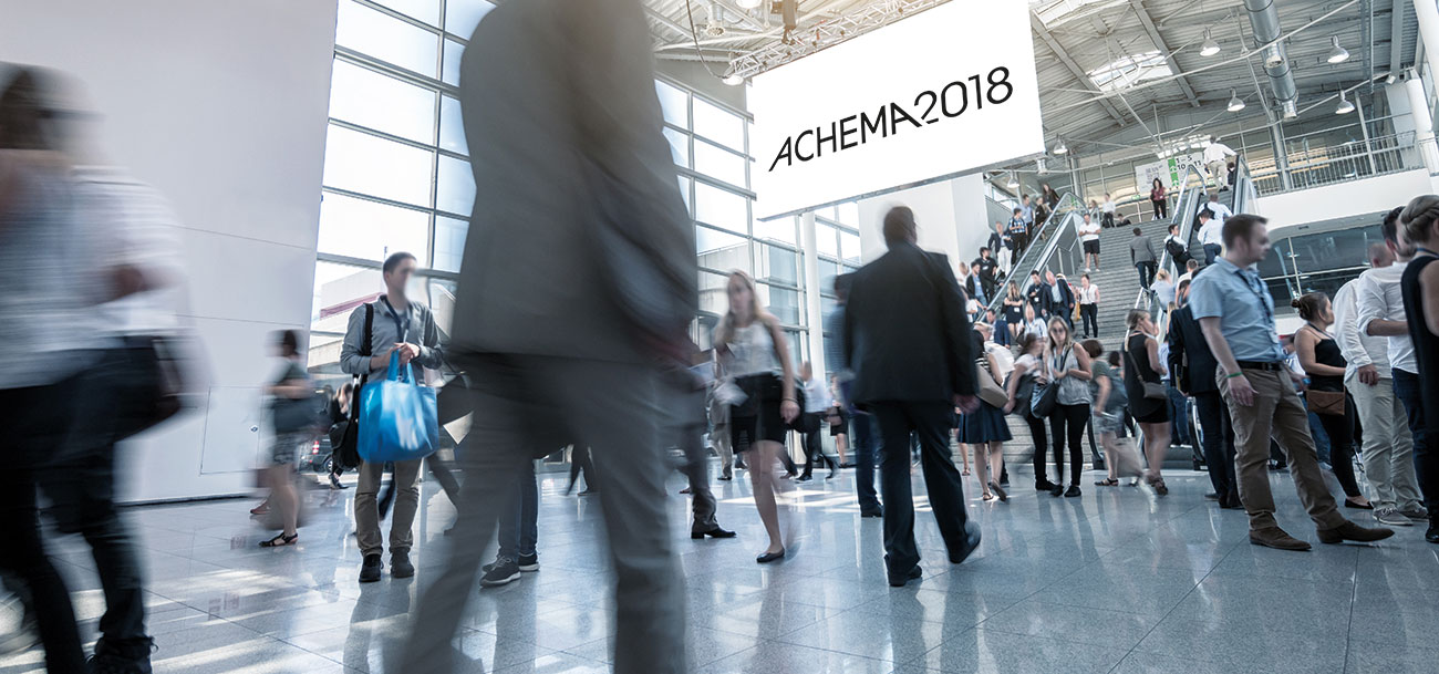 Interior view of ACHEMA 2018 trade fair showing crowds of visitors in a modern exhibition hall with high ceilings, glass walls, and industrial design elements