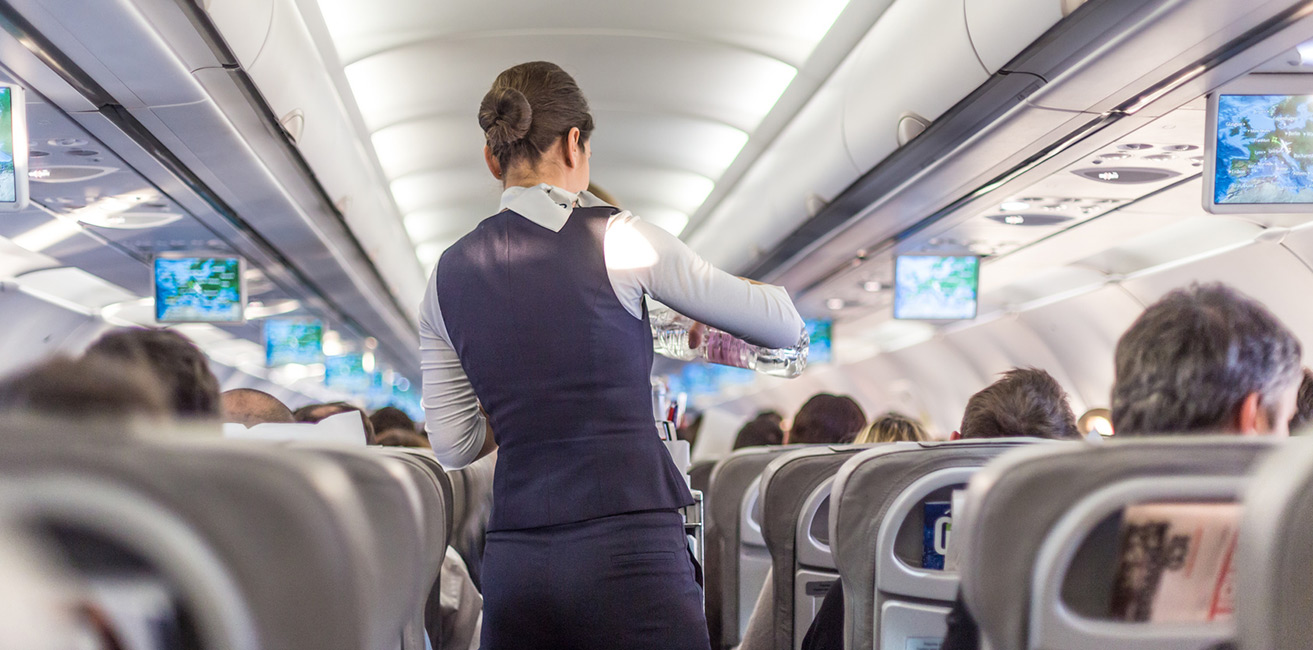 Flugbegleiterin in Uniform geht durch den Gang eines Passagierflugzeugs während des Service, Blick von hinten auf die Kabine mit besetzten Sitzreihen und Bildschirmen an der Decke